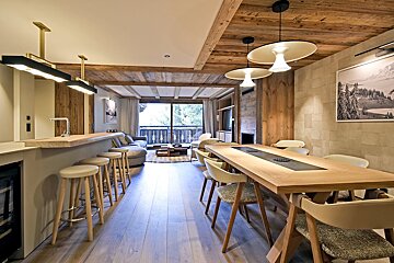 Modern chalet interior featuring a long wooden dining table, breakfast bar with stools, wood-paneled ceilings, and warm, stylish lighting.
