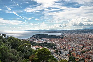 A panoramic view of a coastal city with a large bay, green hills, and a blue sky streaked with airplane contrails.