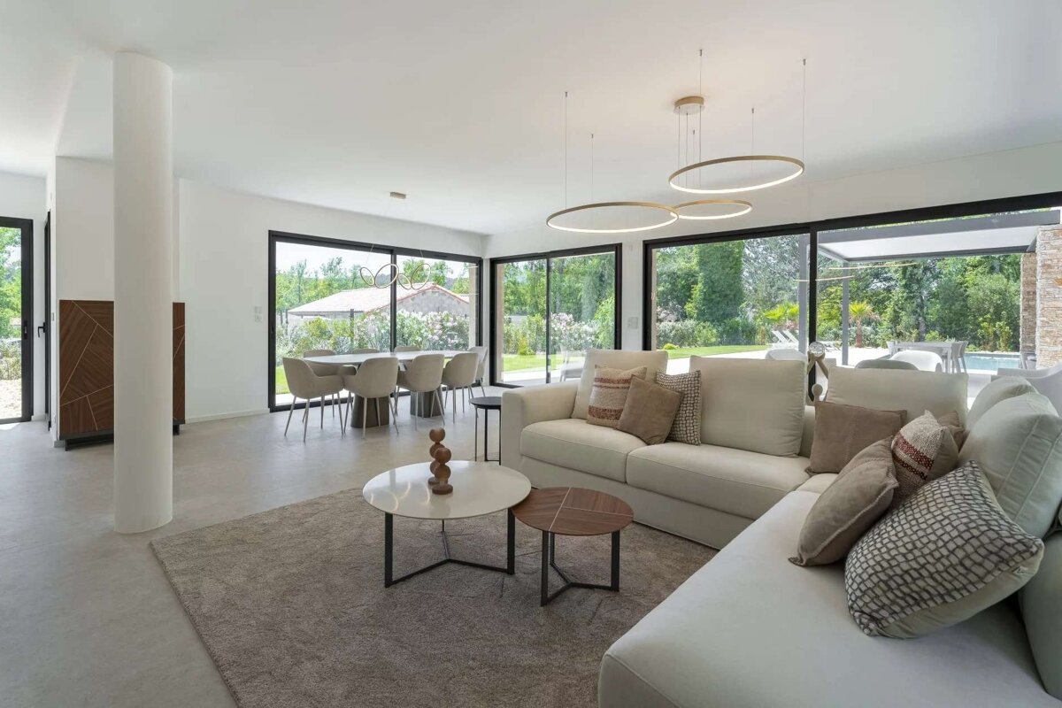 Modern, open-concept living and dining room with large windows overlooking a lush garden and pool, featuring neutral tones and unique circular lights.