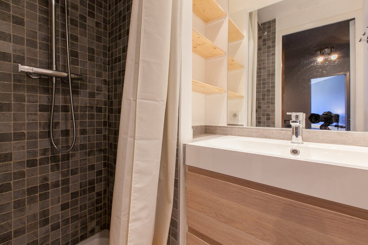 Modern bathroom with dark tiled shower, white curtain, vanity with white sink, wood cabinets, and mirror reflecting shelves and a camera.