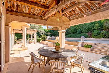A sunny Mediterranean patio featuring a round dining table under a wooden ceiling, overlooking an outdoor swimming pool and garden.