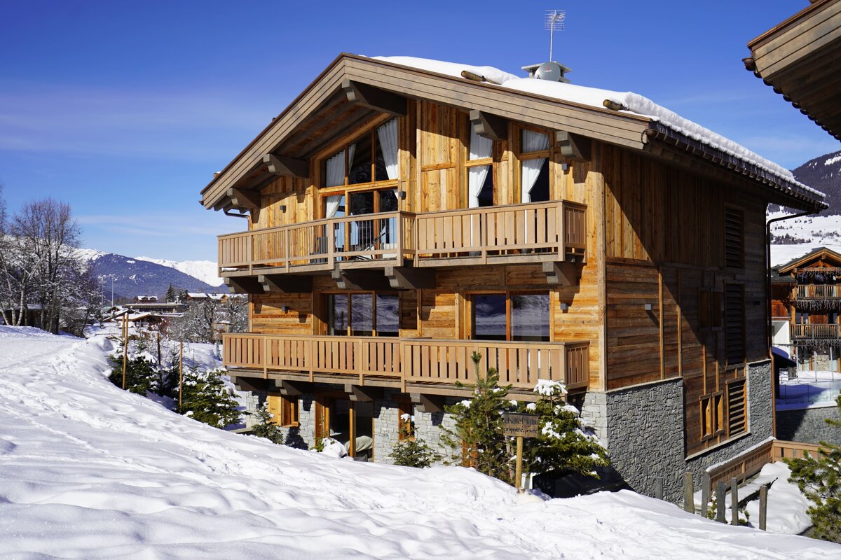 A wooden chalet with balconies stands in a snowy mountain landscape under a clear blue sky.