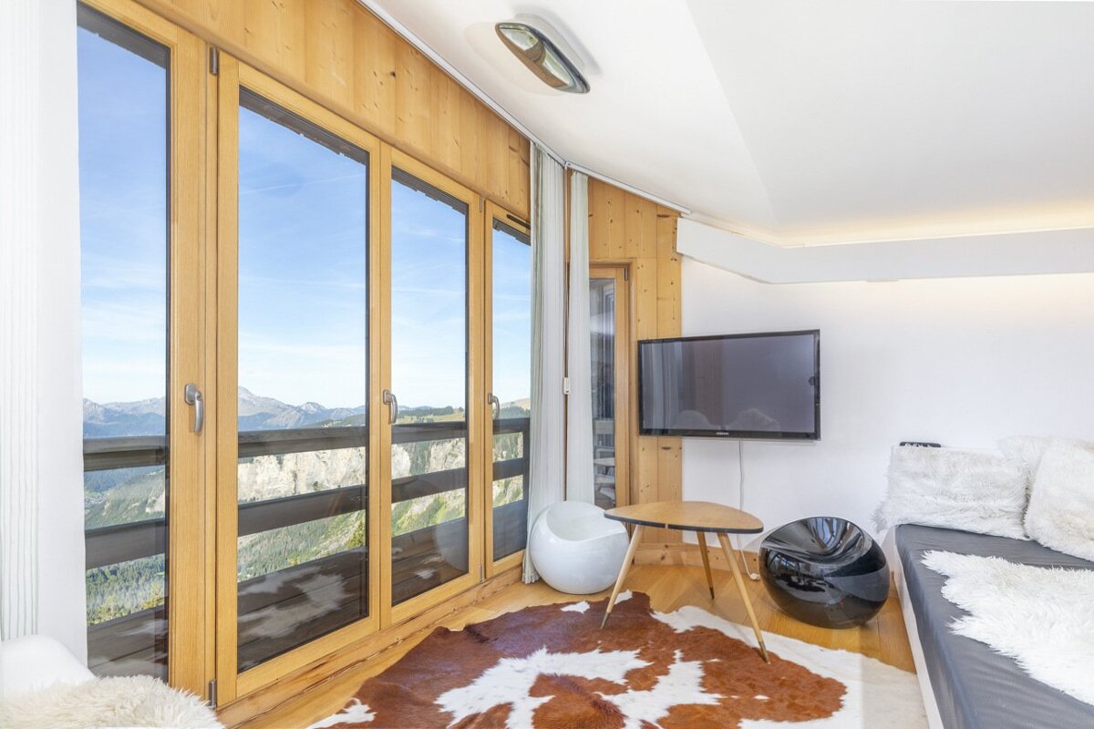 Modern cabin living room with a cowhide rug, sleek chairs, and large wooden windows looking out at mountain peaks.