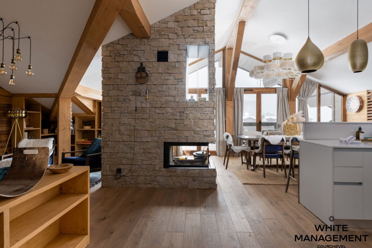 Modern rustic chalet interior with exposed wood beams, a central stone fireplace, light wood floors, and a dining area in the background.