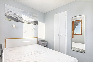 Bright, minimalist bedroom with light blue walls, a white bed, mountain artwork, a white closet, and a full-length mirror reflecting a window.