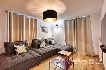 A modern living room features a grey modular sofa, light wood flooring, cream curtains with subtle top lighting, and contemporary ceiling/wall lamps.