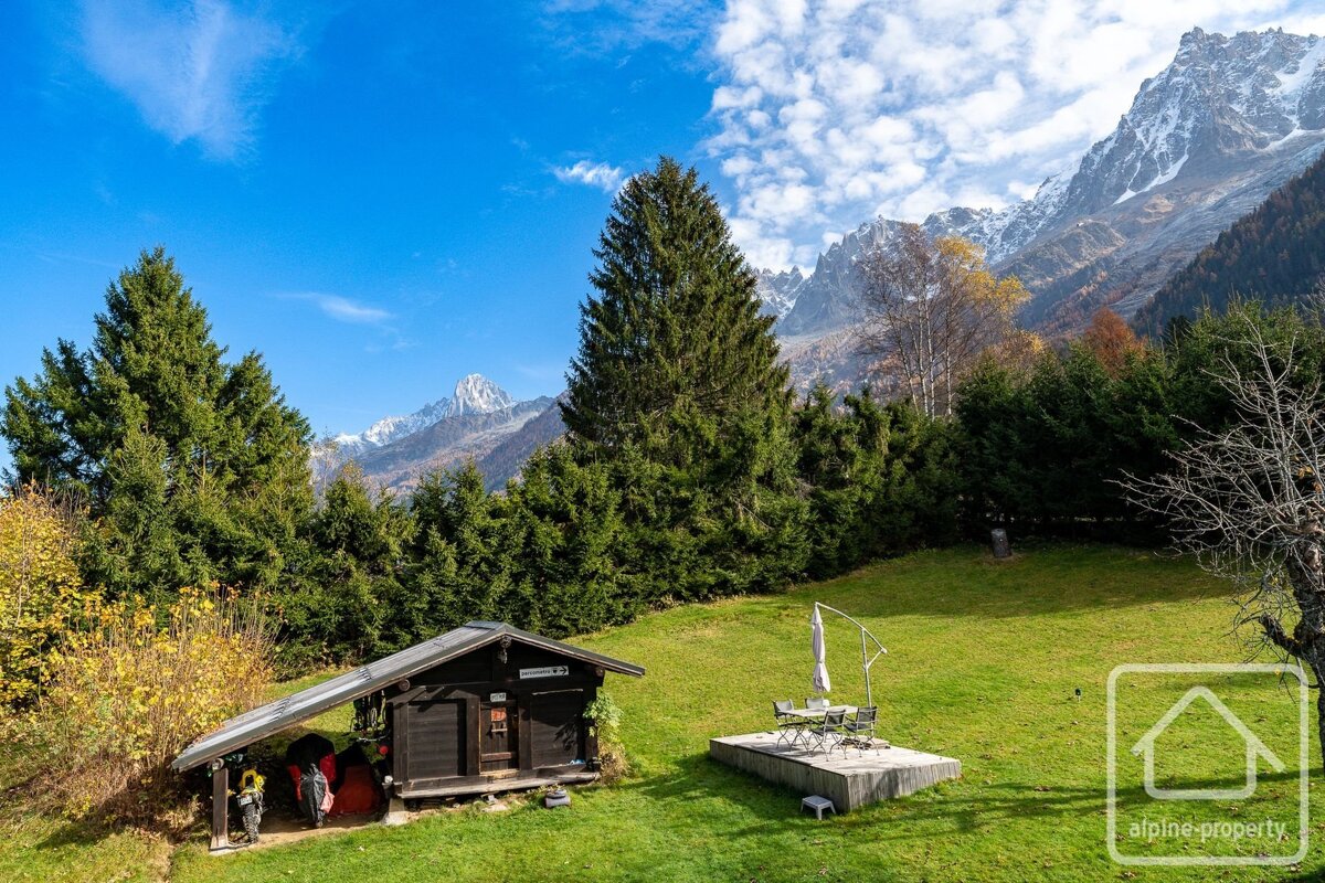 A wooden chalet on a green lawn with a small deck, surrounded by evergreen trees and snow-capped mountains under a blue sky.