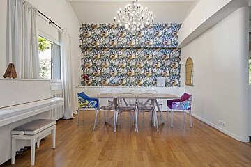 Bright, modern dining room with a long table, clear chairs, colorful armchairs, and floral wallpaper. A white piano and chandelier complete the stylish space.