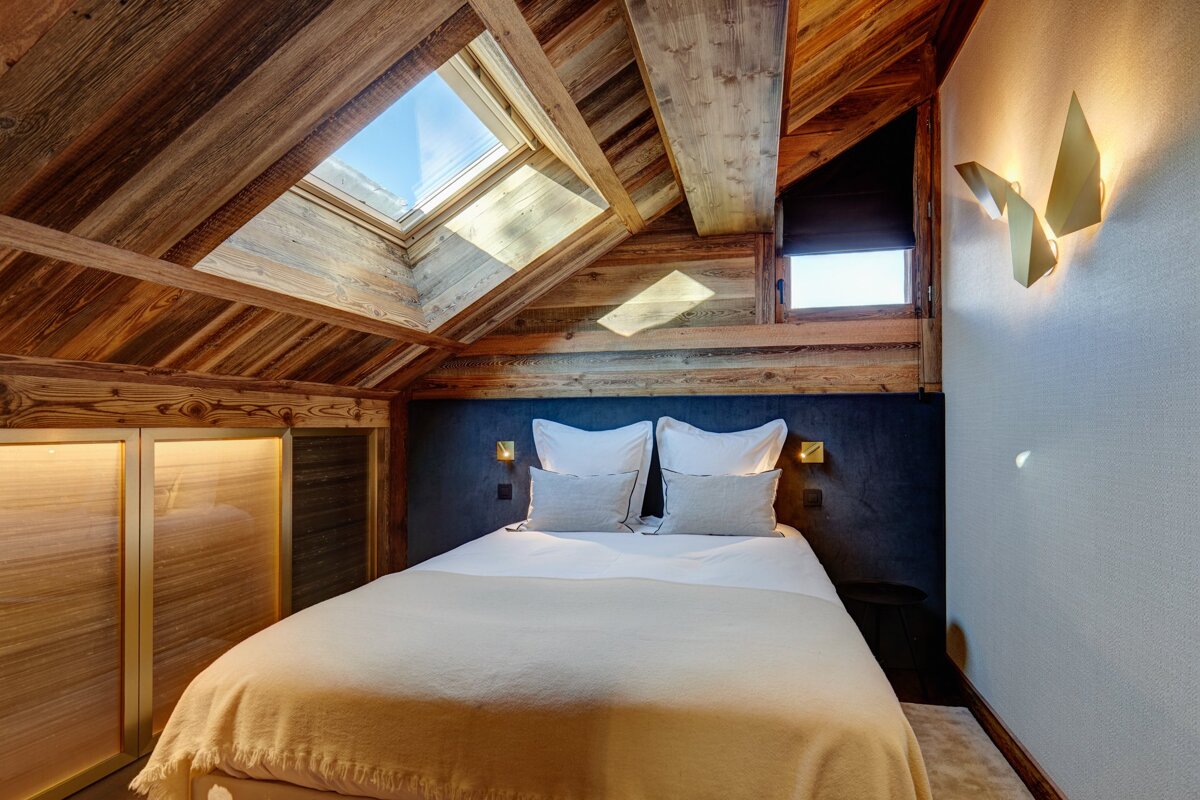 A cozy, rustic-chic bedroom with a slanted wooden ceiling, skylight, large bed, and modern accent lighting in a chalet style.