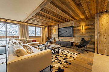 Cozy chalet living room with wood walls and ceiling, a large beige sofa, a unique patterned rug, and snowy mountain views.