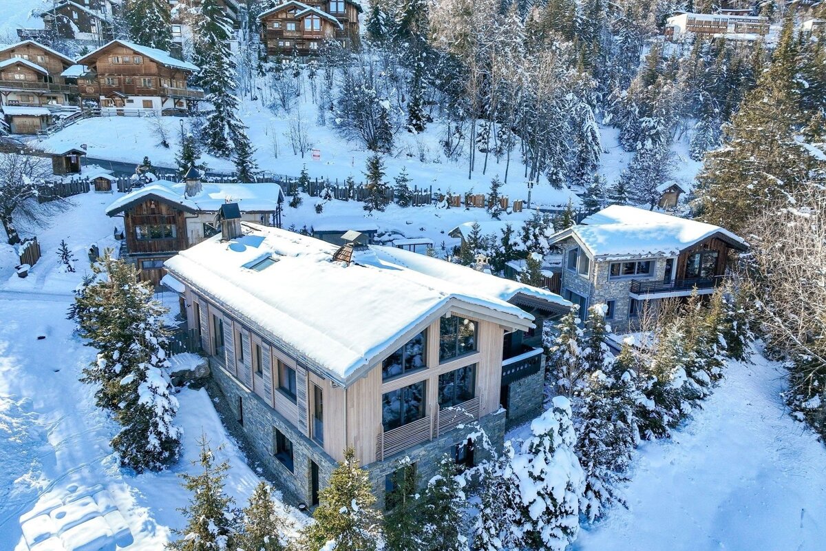 An aerial view of a picturesque snowy mountain village with traditional chalets nestled among snow-covered evergreen trees under a clear sky.