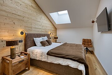 A modern, cozy attic bedroom featuring a wood-paneled wall, brown bed, skylight, and warm lighting. Includes a wooden nightstand and wall-mounted TV.