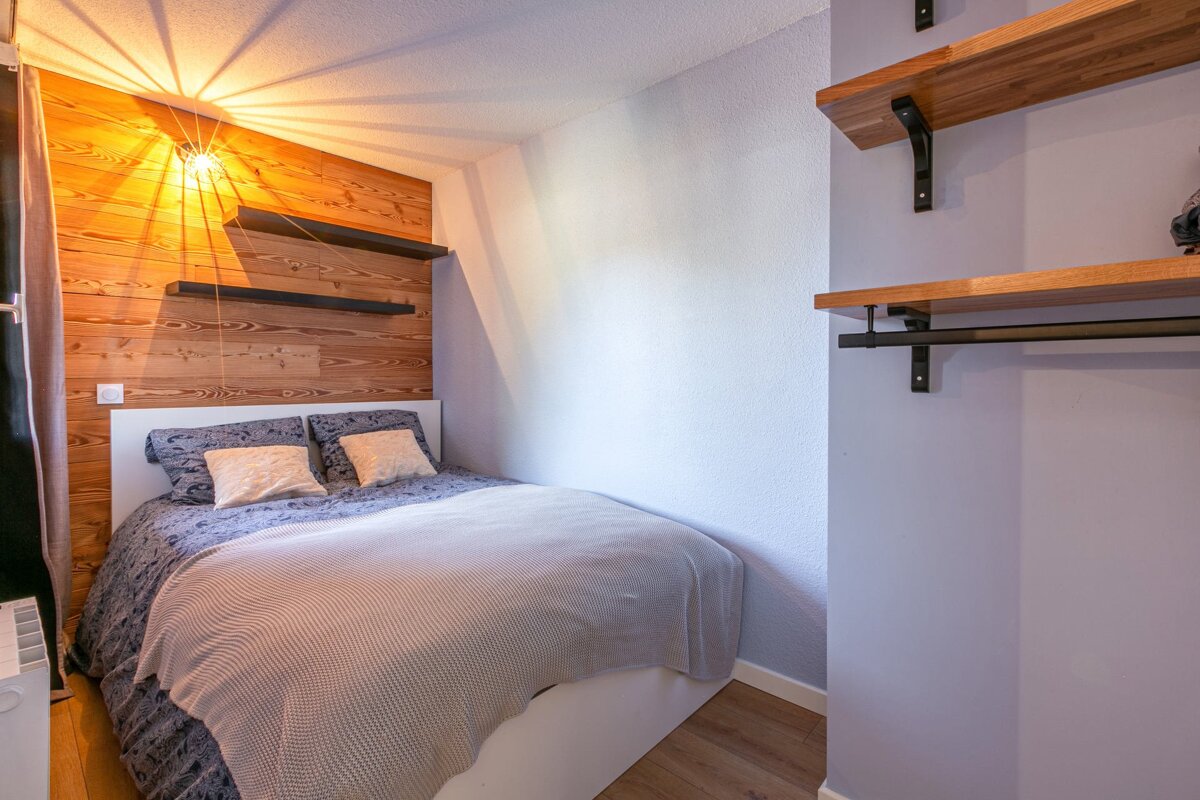 A cozy bedroom features a bed with patterned bedding against a wooden accent wall with shelves. A bright light illuminates the ceiling.