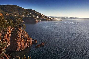 Vast blue sea and red rocky coast with a hillside town under a clear sky, fading into a hazy distant horizon.