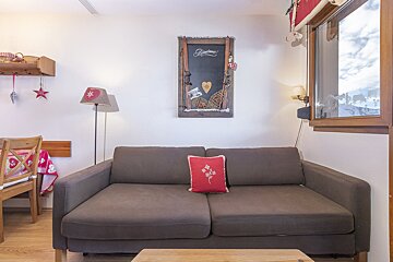 A cozy, chalet-style living room with a brown sofa, alpine decor including a ski-themed sign, and a window revealing a snowy mountain view.