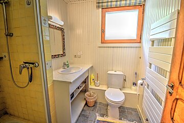 A clean bathroom with a yellow tiled shower, white paneled walls, a vanity with sink, toilet, and a window. Gray floor tiles.