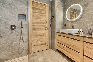 Modern bathroom featuring grey stone tiles, a light wood door and vanity, a vessel sink, and a circular backlit mirror.