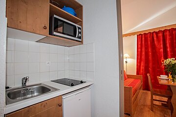 A small apartment kitchen with a sink, microwave, stovetop, and mini-fridge, adjacent to a living area with red curtains, a dining table, and a sofa.