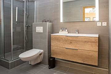 Modern gray-tiled bathroom with a glass shower, wall-mounted toilet, and a wooden vanity under an illuminated mirror.
