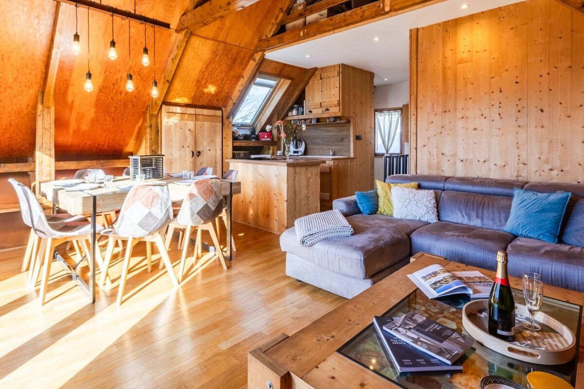 A cozy, sunlit wooden chalet interior featuring a living area with a sofa, a dining table, and a rustic kitchen.