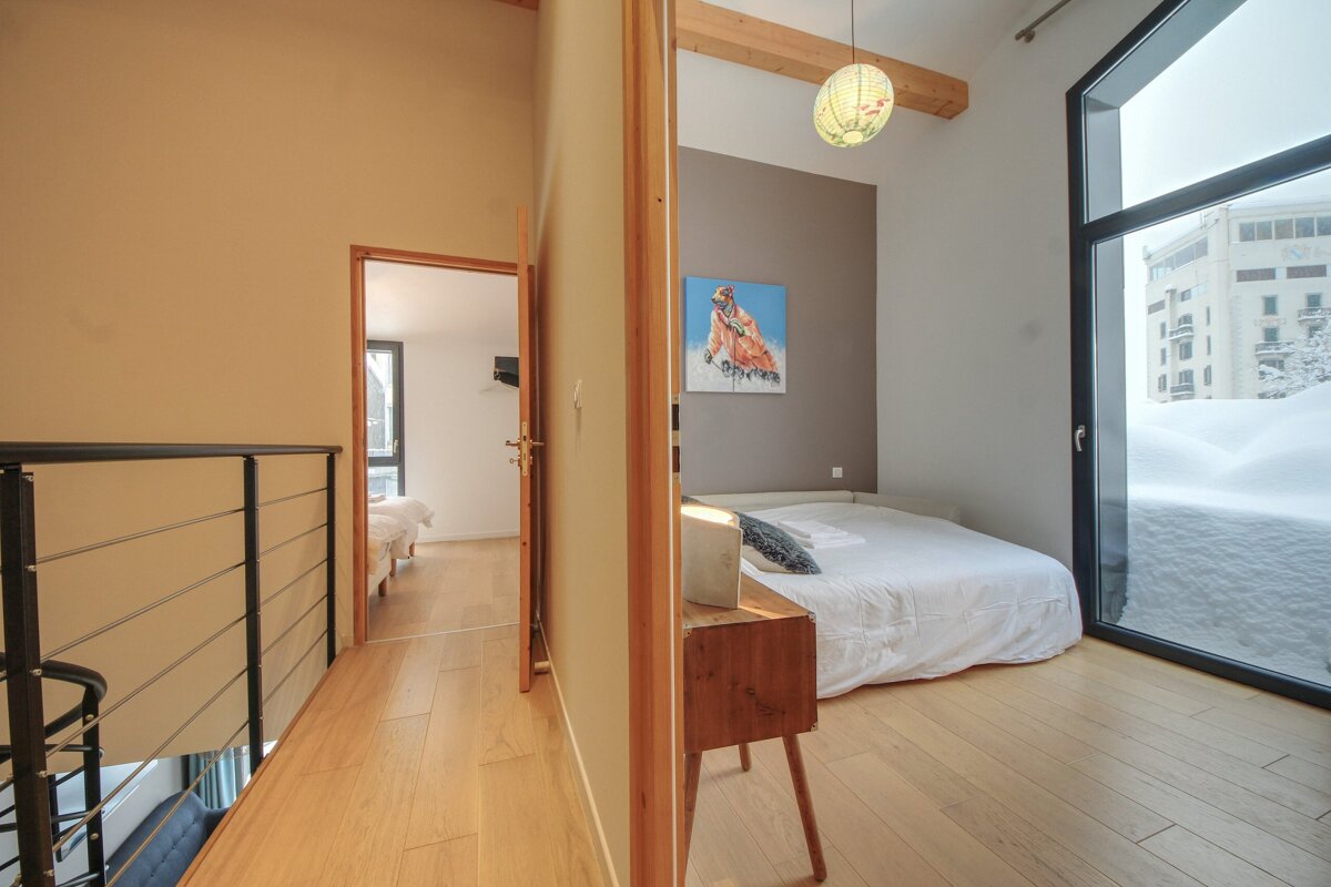 A cozy bedroom with a white bed, abstract art, and a large window showcasing deep snow. Adjacent is a loft space with a railing and another room visible.