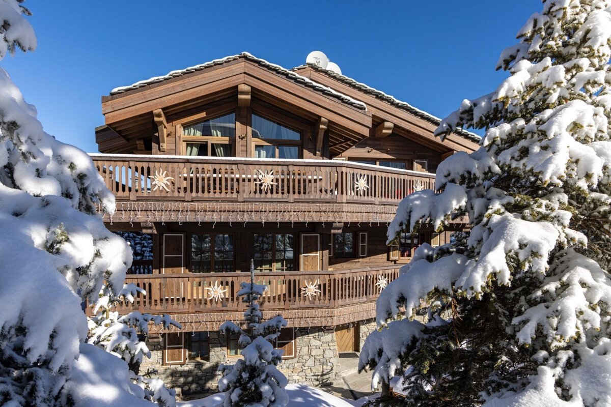 A snow-covered wooden chalet with balconies and evergreen trees under a clear blue sky, evoking a cozy winter scene.