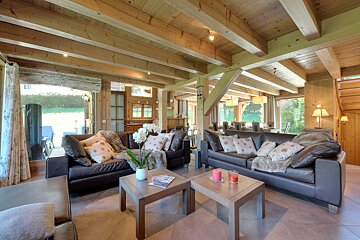 A bright, open-plan living space with exposed wooden beams, dark leather sofas, and large windows. Rustic modern design, connected to dining and outdoors.