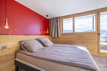 Cozy alpine bedroom with a bold red accent wall, wood paneling, a grey double bed, and large windows showing snowy mountain views.