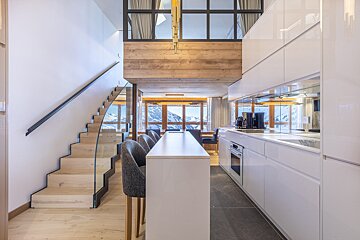 Luxurious ski chalet interior featuring a sleek white kitchen, elegant wooden stairs, and a stunning view of snow-capped mountains.