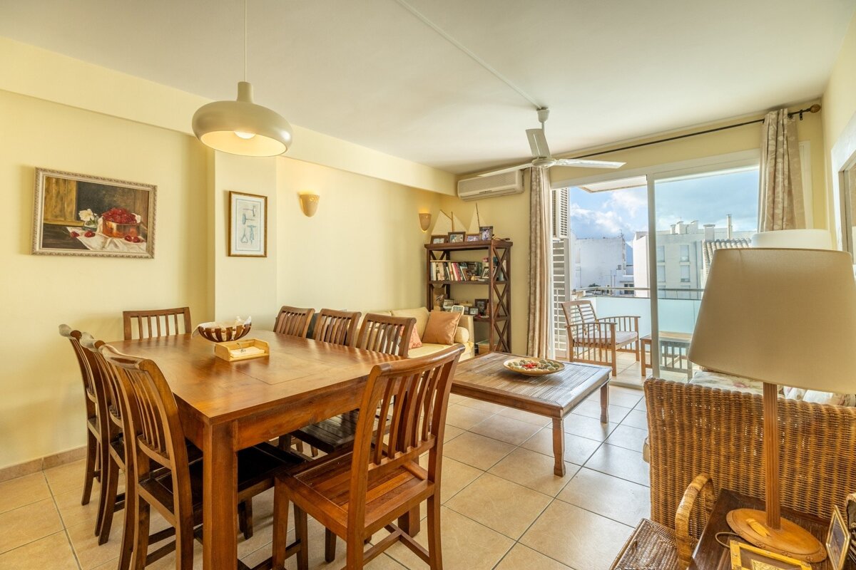 A bright, open-plan living and dining area featuring a wooden dining table, comfy seating, and balcony access, with warm yellow walls and tiled floors.