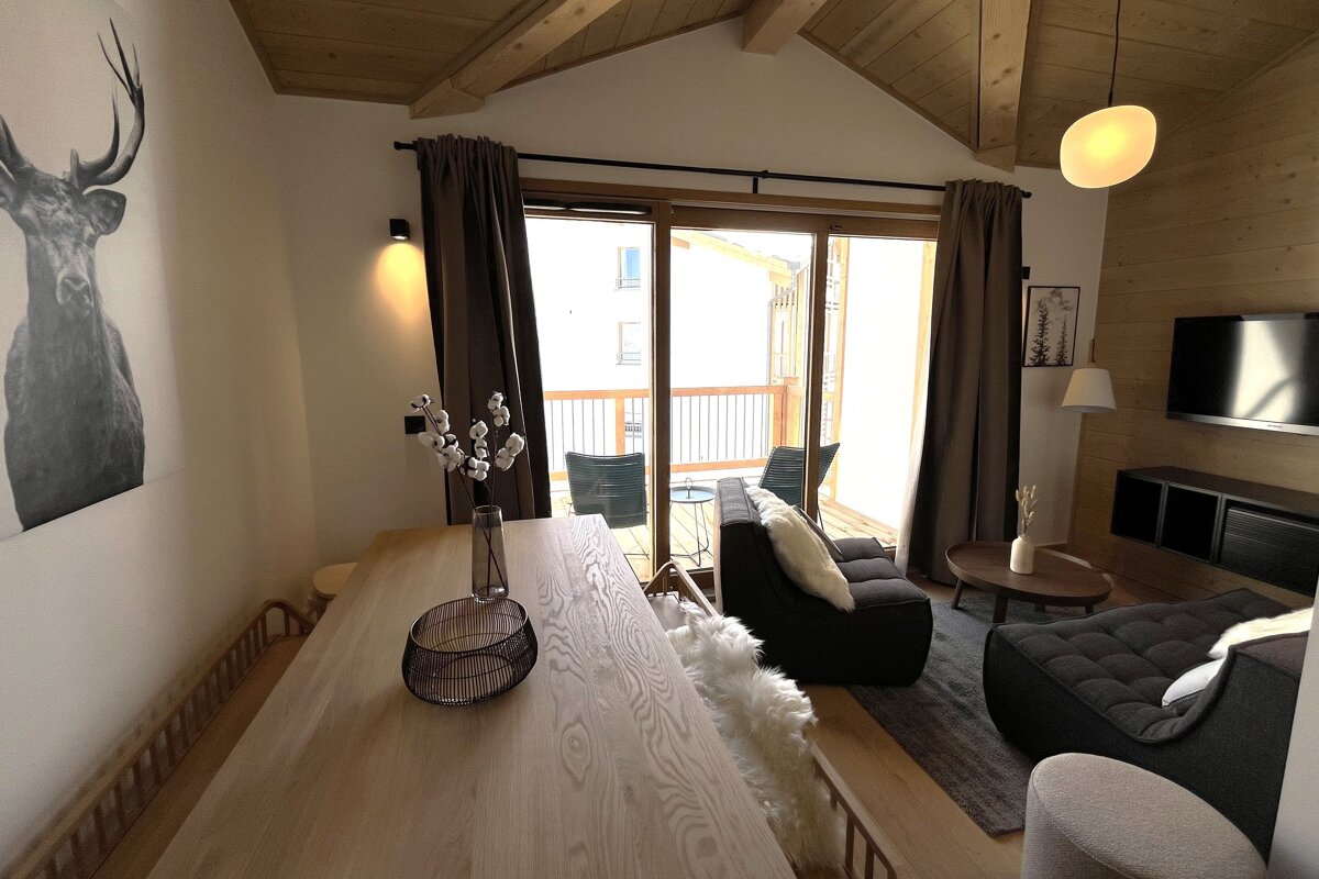 A modern, cozy alpine living and dining room with a wooden vaulted ceiling, stag art, comfy seating, and large glass doors leading to a balcony.