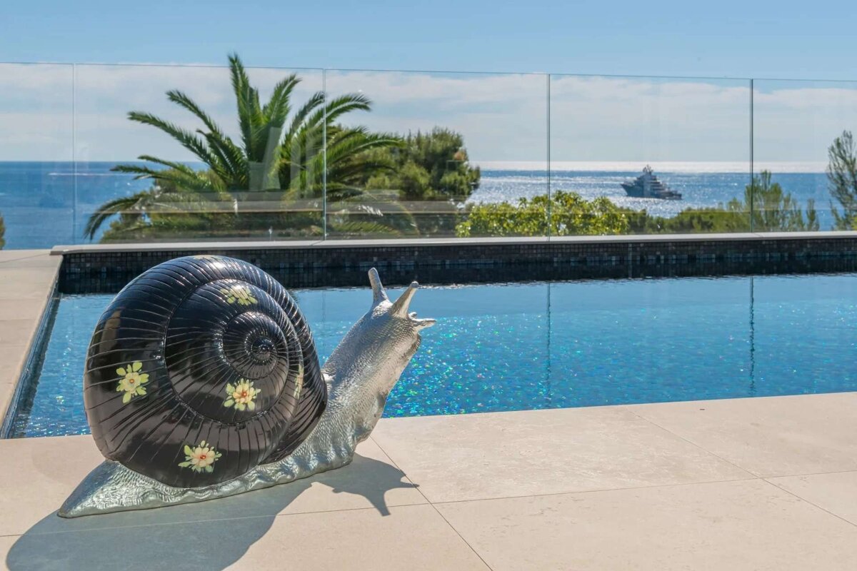 A large, black and silver snail sculpture by a sparkling pool overlooks the ocean with a yacht and palm trees under a clear sky.