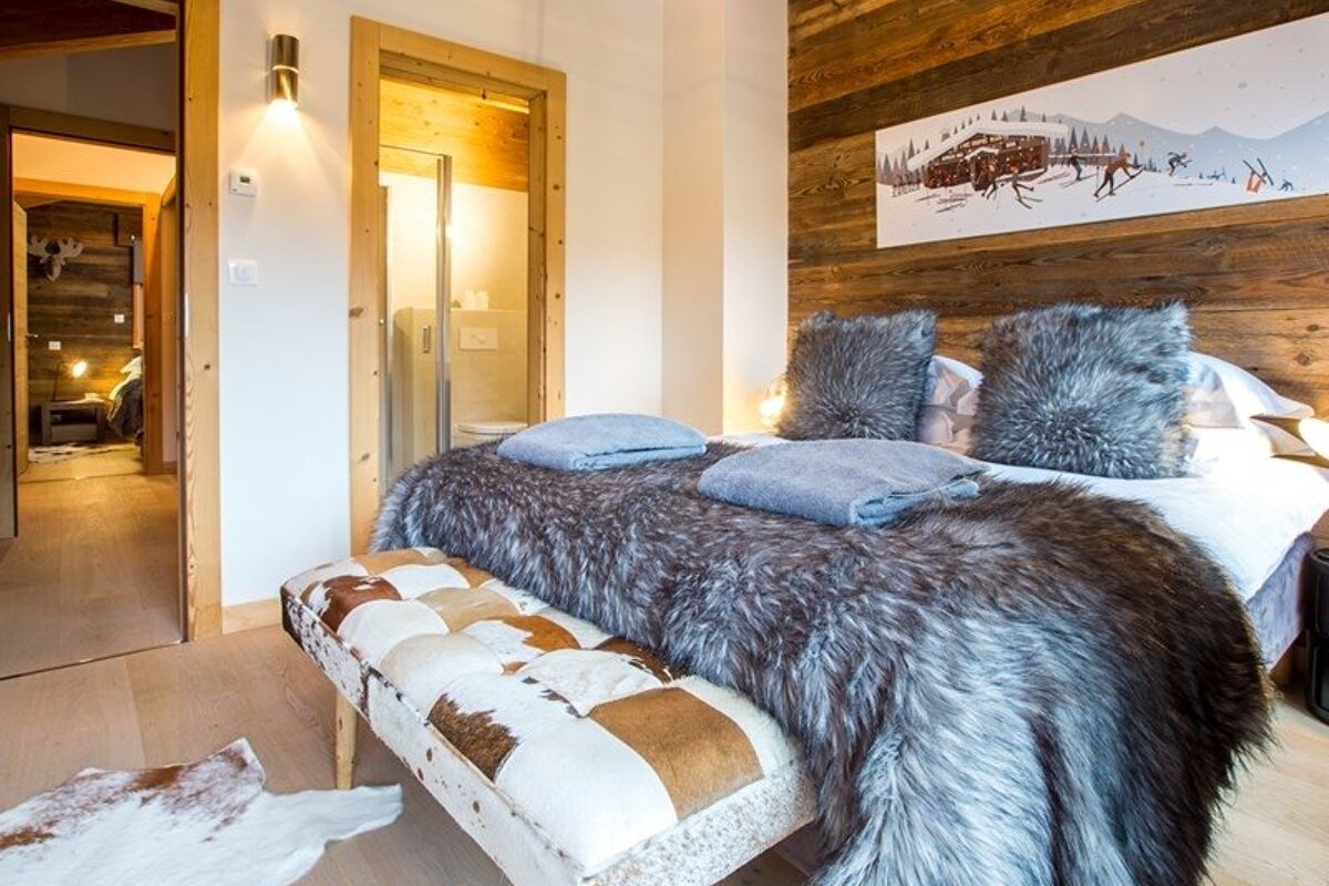 Cozy chalet bedroom with a plush fur blanket, furry pillows, a cowhide bench, and rustic wood-paneled walls.