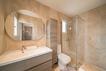 A beige-tiled bathroom featuring a floating sink vanity with a large round mirror, a toilet, and a glass-enclosed shower.