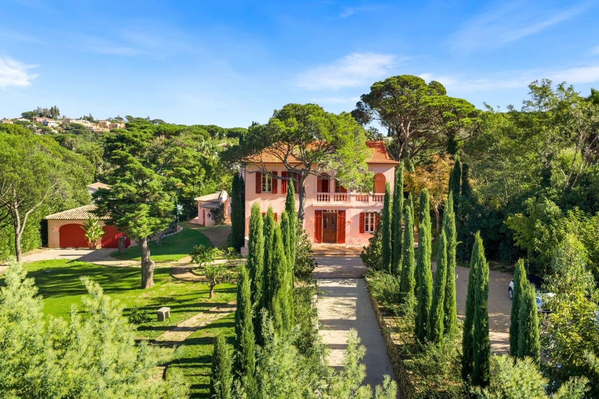 A sunny pink Mediterranean villa surrounded by lush lawns, tall cypress trees, and green foliage under a clear blue sky.