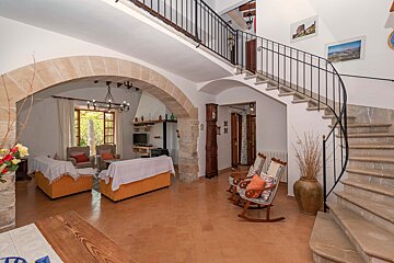 A spacious rustic interior with stone arches, a living area, terracotta floor, and a grand curved staircase leading to an upper floor.