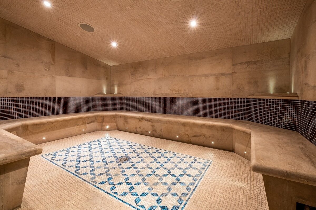 A luxurious spa room with light brown tiled walls, built-in benches, and a blue and white geometric mosaic floor, illuminated by soft lighting.
