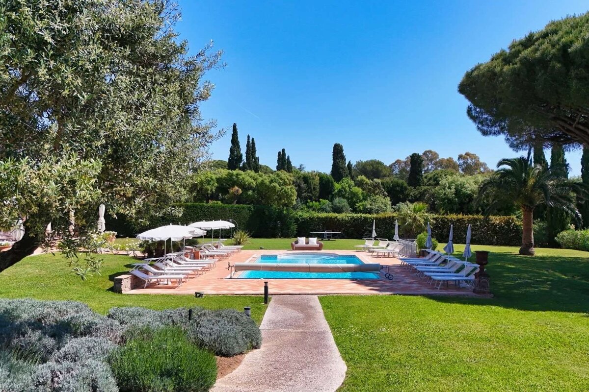 Lush green garden with a sparkling blue pool, numerous lounge chairs, white umbrellas, and tall trees under a clear blue sky.