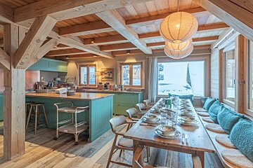 Cozy chalet dining room and kitchen with exposed wooden beams, teal accents, and a large window looking out onto a snowy winter scene.
