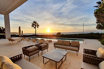 The sun is setting over the ocean behind a couch and chairs