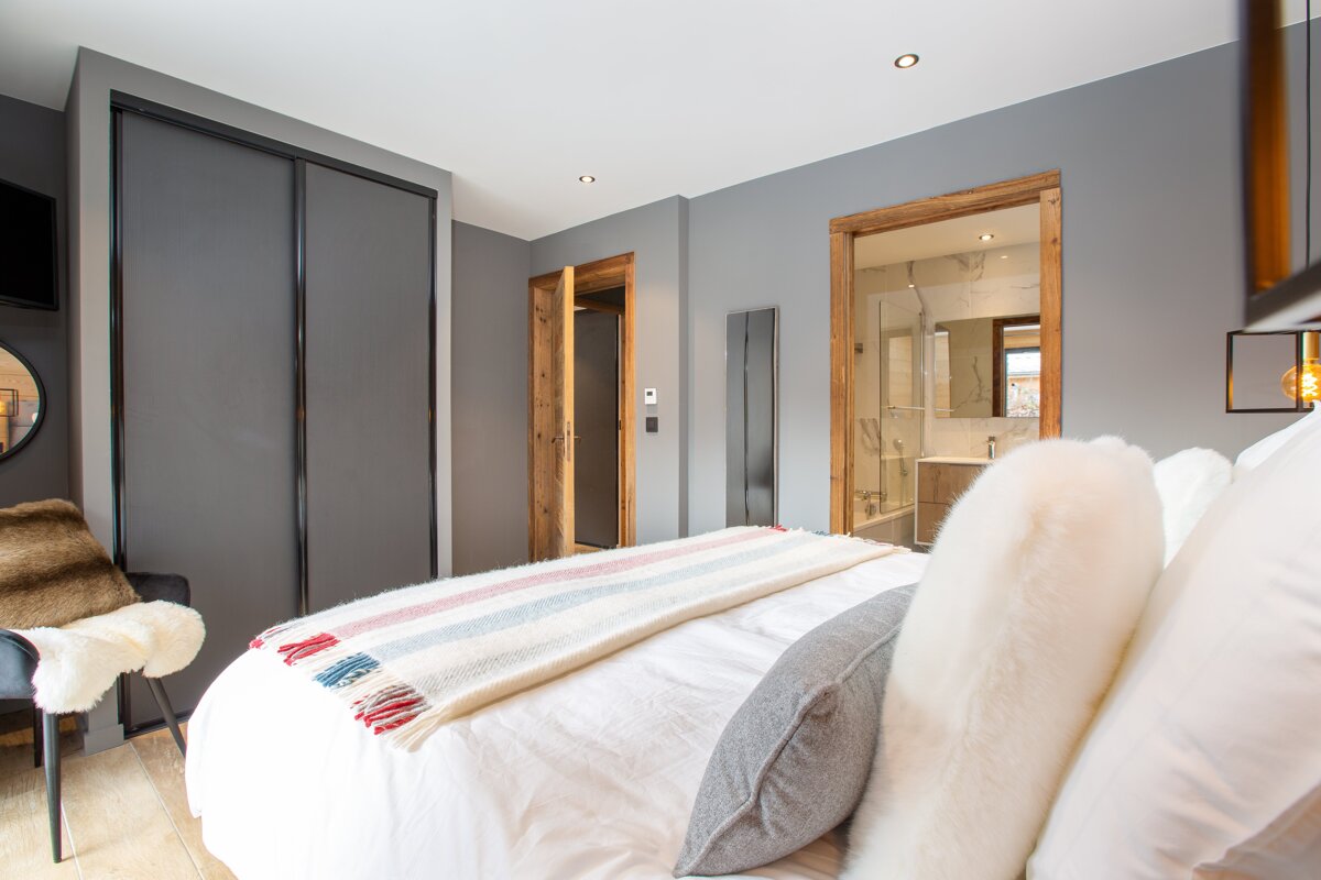 A modern gray bedroom with white bedding, a colorful striped throw, and wooden doorframes. An en-suite bathroom is visible, and sleek wardrobes are present.