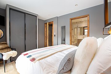 A modern gray bedroom with white bedding, a colorful striped throw, and wooden doorframes. An en-suite bathroom is visible, and sleek wardrobes are present.