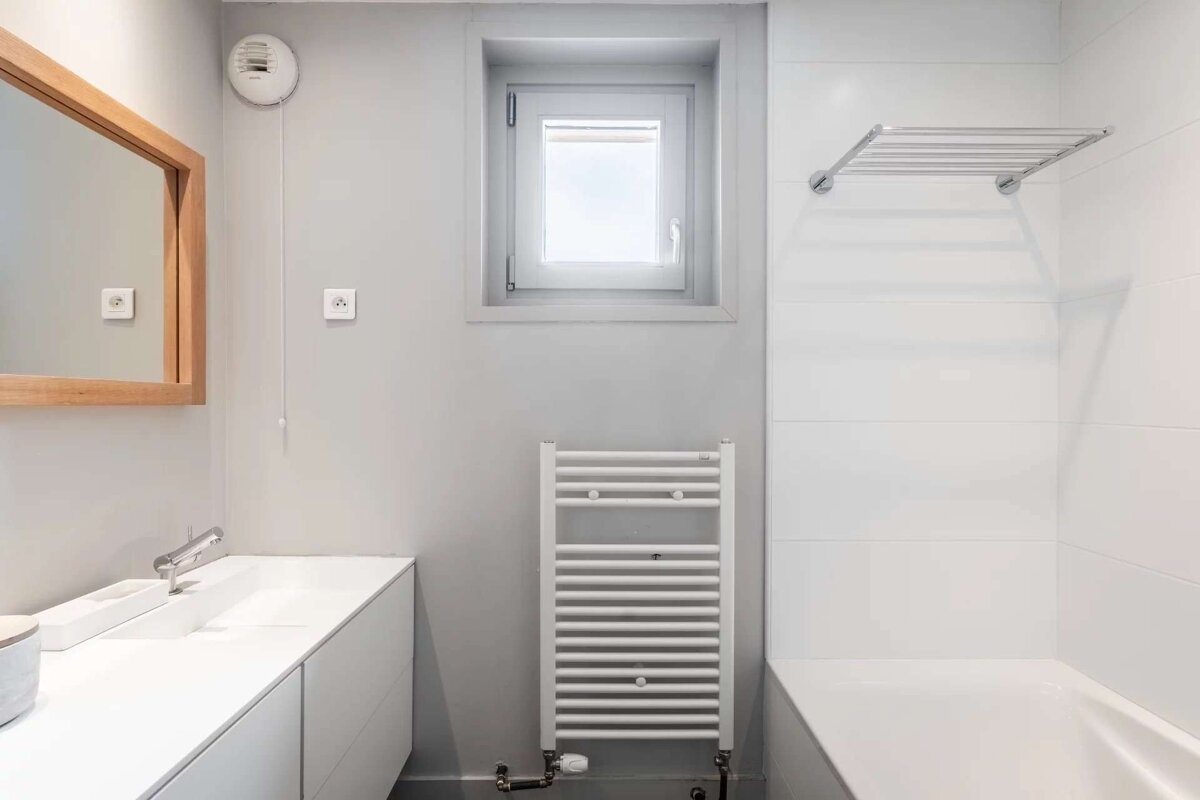 A modern bathroom with a white vanity, wood-framed mirror, small window, towel warmer, bathtub with white tiles, and light gray walls.