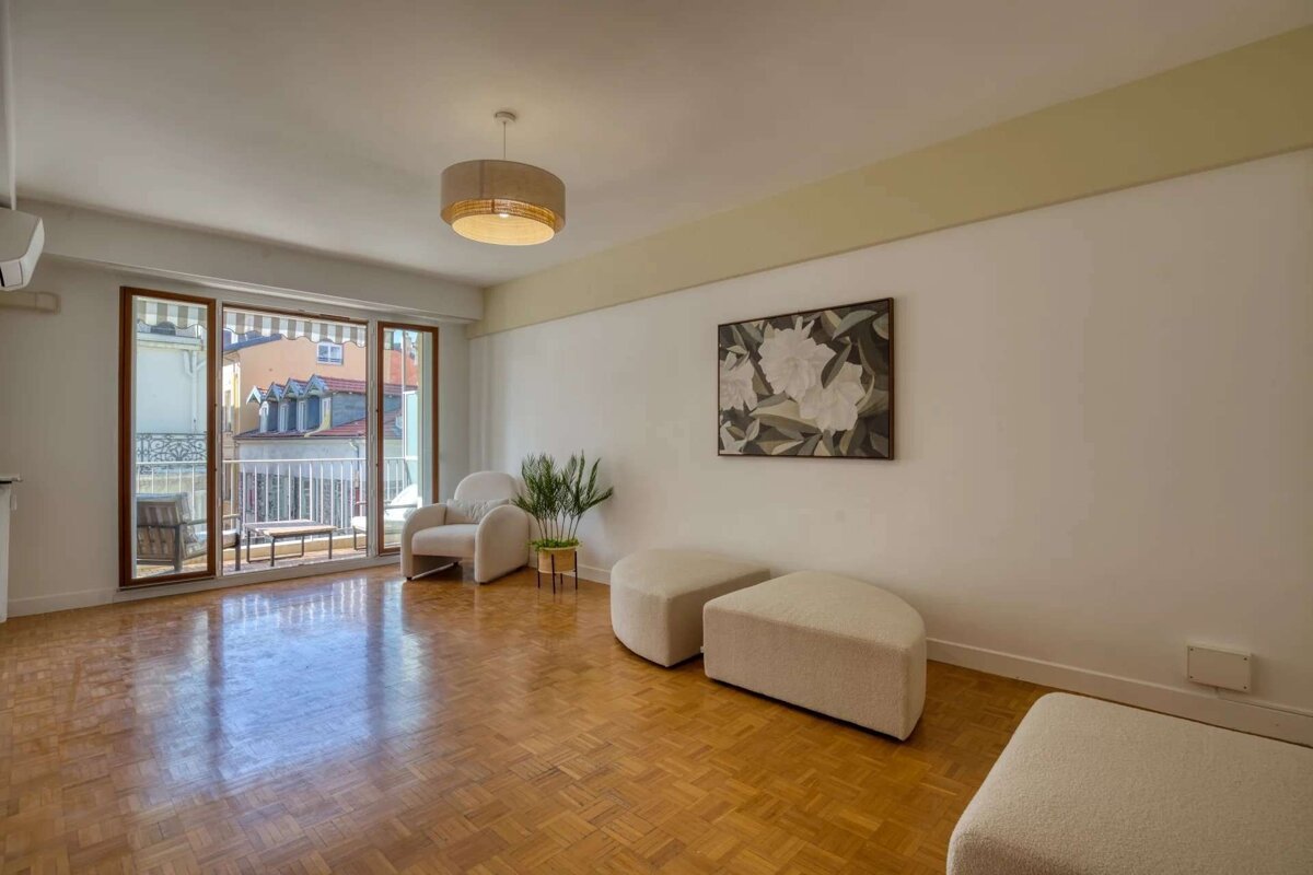 Bright living room with parquet flooring, balcony, white armchair, neutral ottomans, plant, and floral art on white walls.