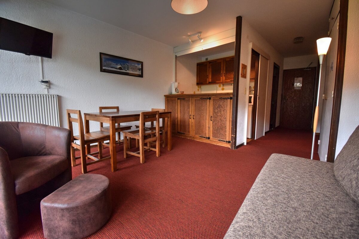 A cozy living area with a red carpet, wooden dining table, kitchenette, TV, sofa, and armchair, leading to a hallway with doors.