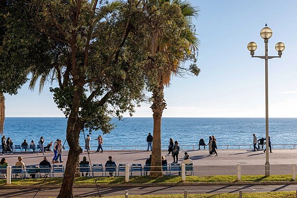 People walk and sit on a sunny seaside promenade with trees and a streetlamp, overlooking a calm blue ocean under a clear sky.