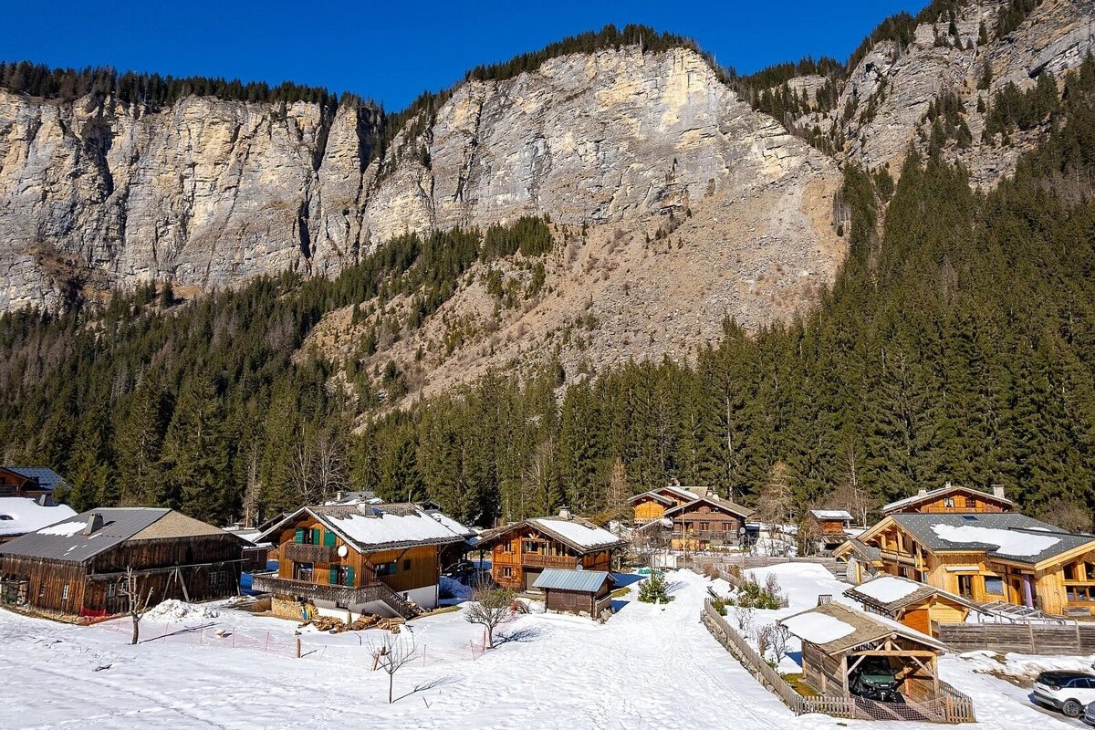 Snowy alpine village with chalets nestled at the base of towering cliffs and a dense pine forest under a clear blue sky.