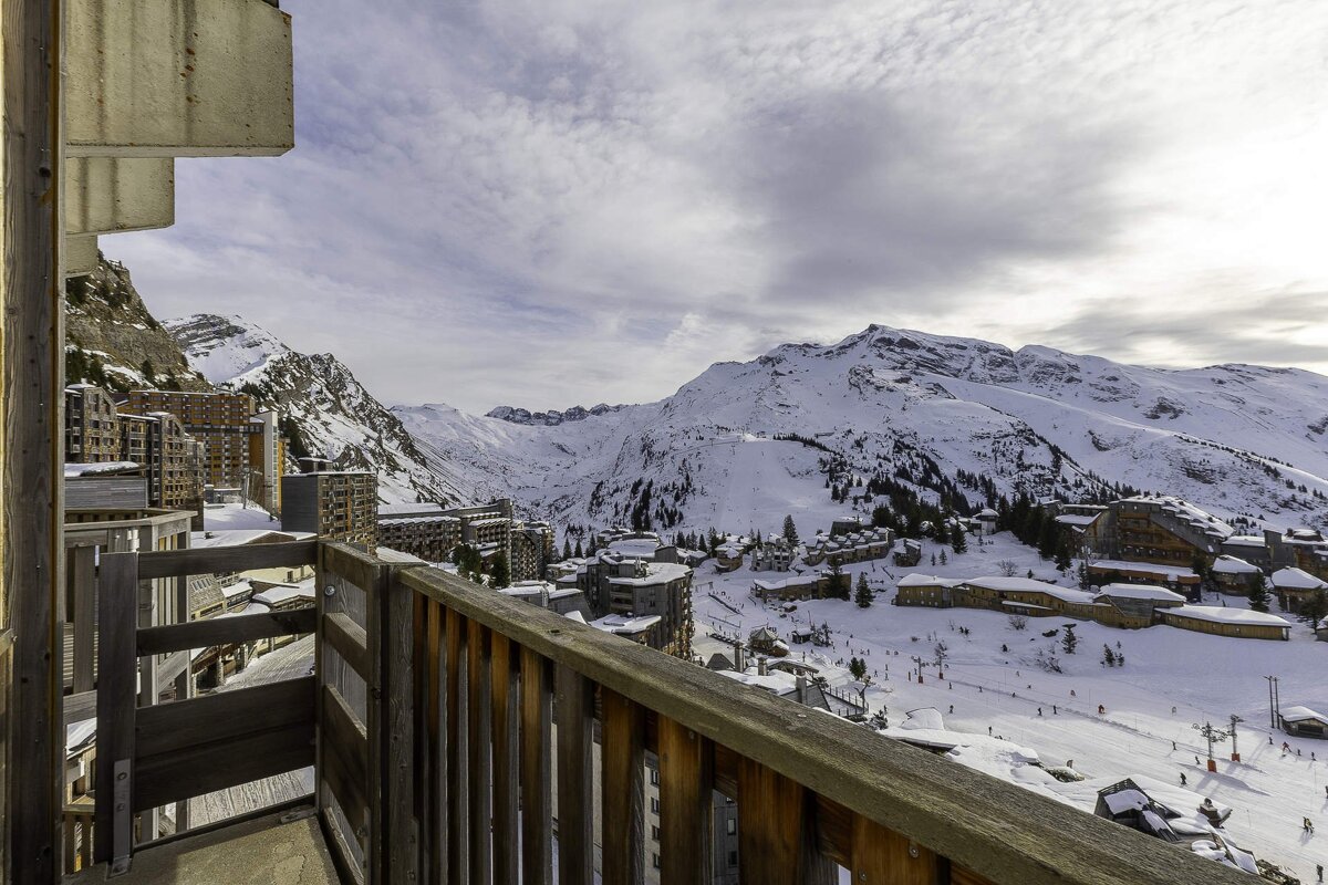 A wooden balcony overlooks a snow-covered mountain ski resort and alpine village under a cloudy sky.