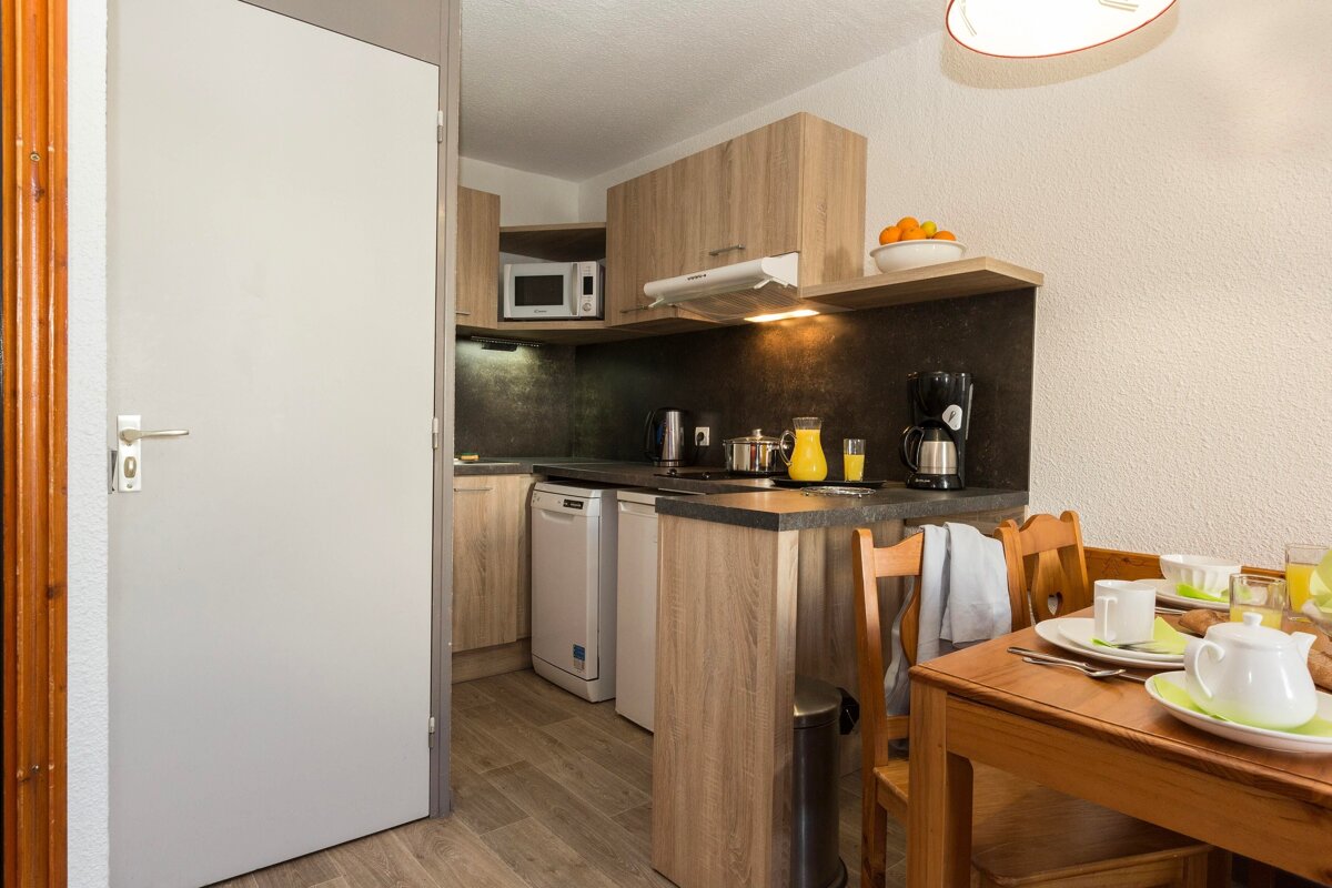 A compact, modern kitchen with wooden cabinets and appliances, next to a dining table set for breakfast, hinting at a cozy mealtime.