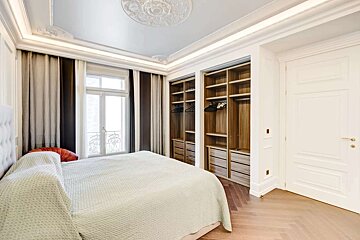 A classic bedroom features a large bed, a window with striped curtains, built-in wooden wardrobes, a white door, and an ornate ceiling with crown molding.